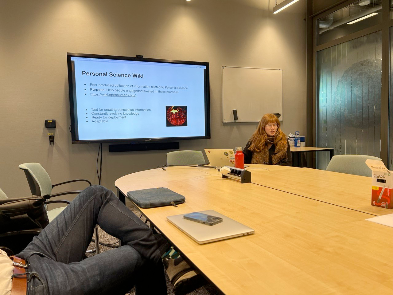 Two people sitting around a conference table, with a screen showing a slide in the background. The slide reads "Personal Science Wiki. in bullet points: 1. Peer-produced collection of information related to personal science 2. Purpose: help people engaged/interested in these practices. 3. Tool for creating consensus information 4. Constantly evolving knowledge 5. Ready for deployment 6. adaptable