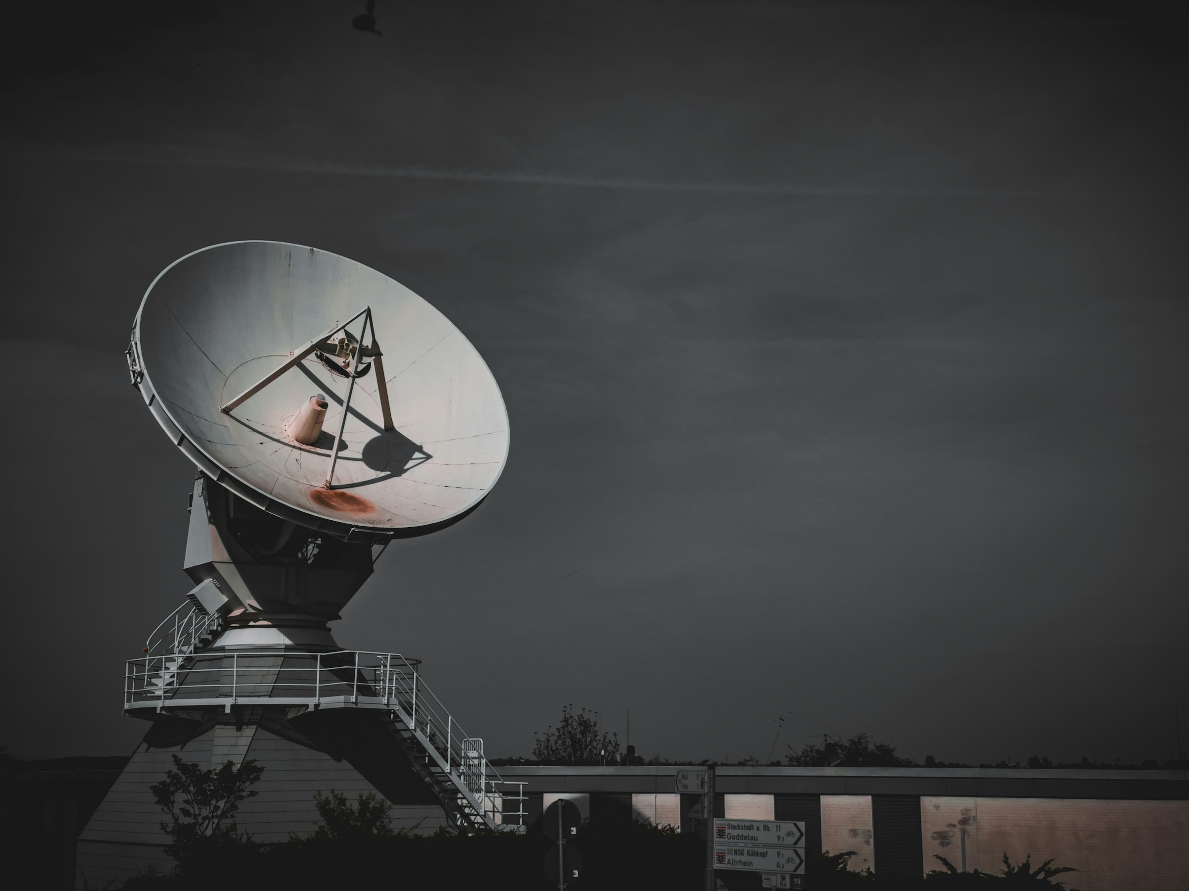 An imposing satellite dish is photographed against a dusky sky, featuring a prominent steel framework and the dish's parabolic shape designed for signal reception and transmission. The scene is composed with the satellite dish dominating the upper half of the frame, underscored by a building with directional signage partially visible below.