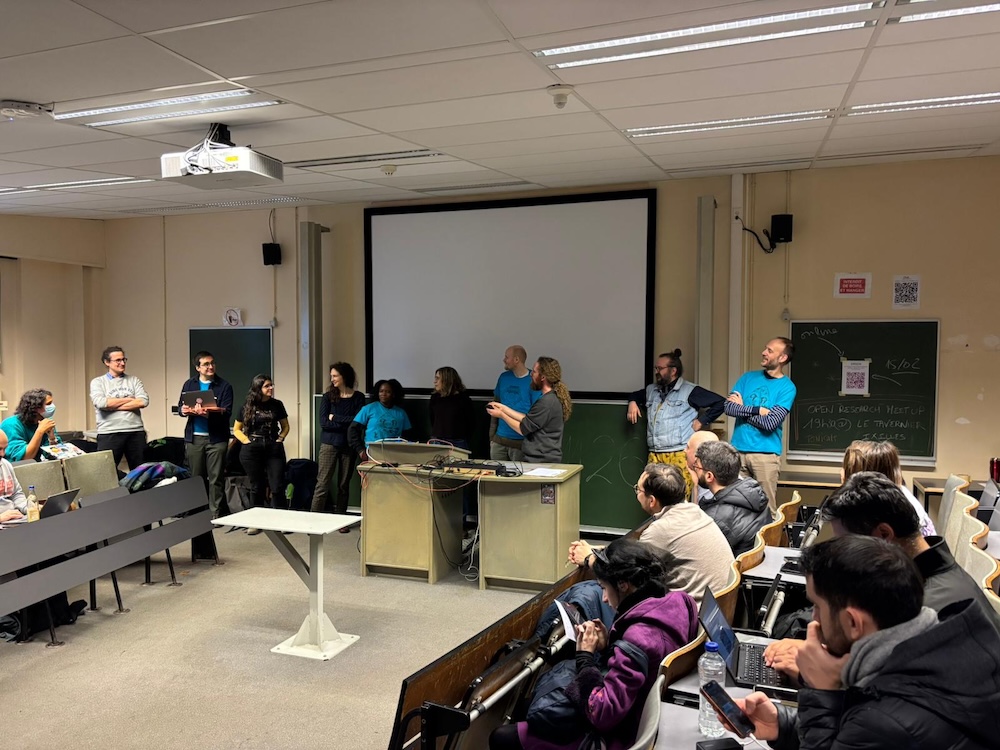 A small classroom, with attendees sitting in rows of chairs, forming an &quot;n&quot; shape. In front of the room is a group of ten (10) people from varying nationalities and gender groups. They are the members of the organising committee of the conference. Behind them is a blank projector screen, and in front, a beige table.