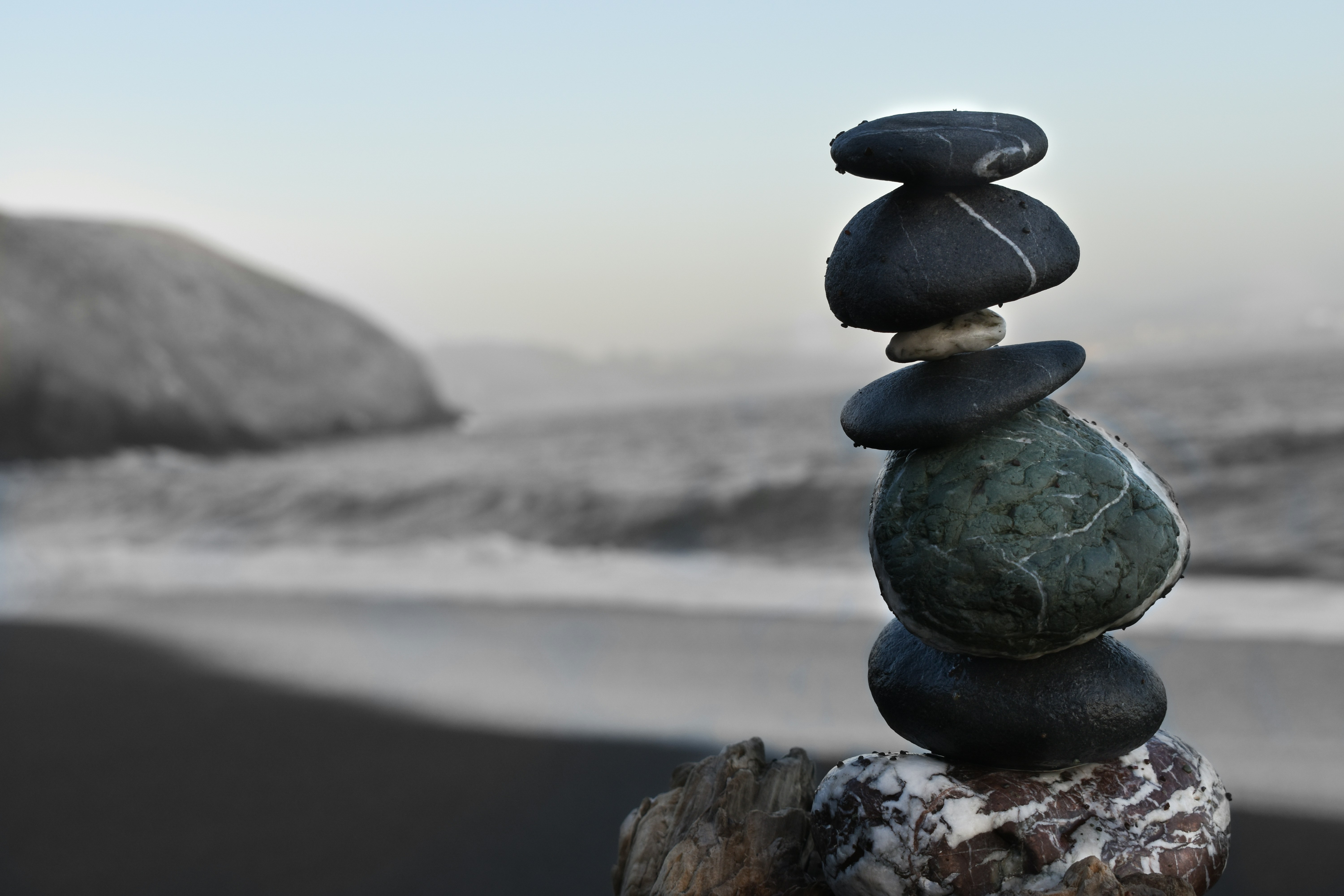 Five black rocks stacked and balanced on top of each other with a beach and waves in the background