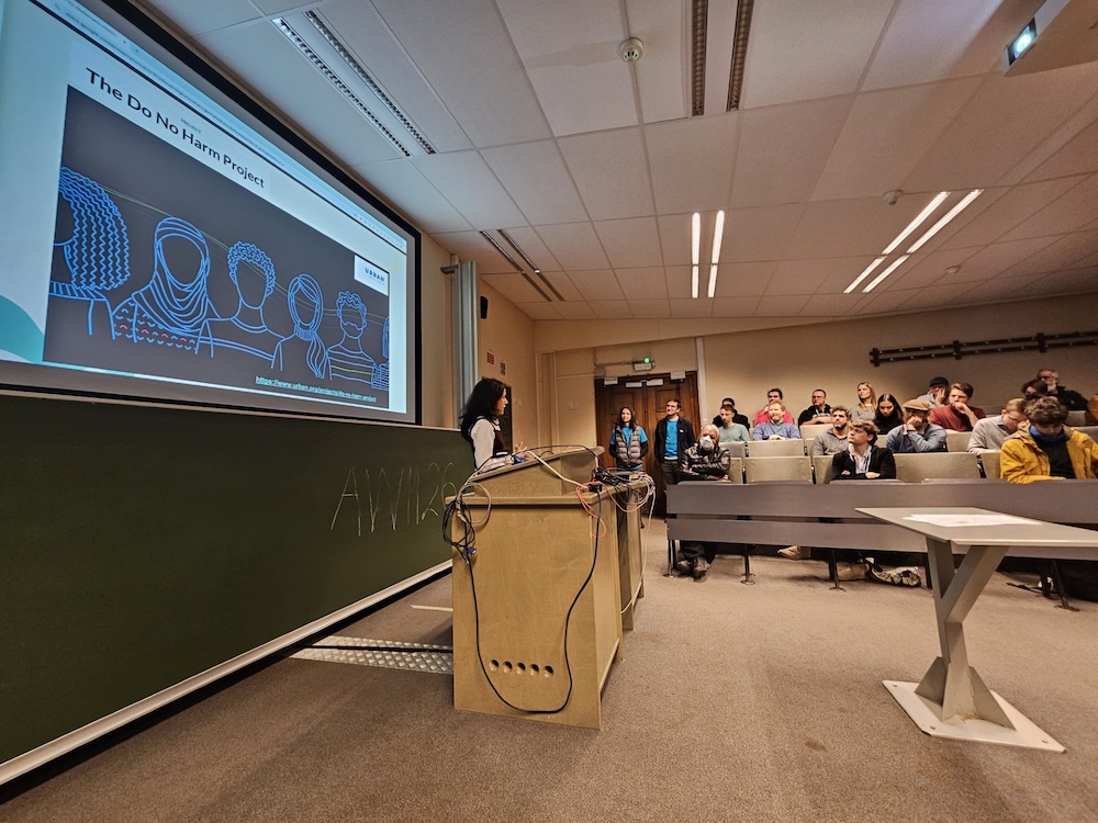 A conference room photographed from the left side of the room, showing a speaker, and some members of the audience. The speaker, Malvika, is an Indian woman of small stature and silky dark hair, wearing a white long-sleeved shirt under a sleeveless maroon dress. She is presenting from a slide that reads The Do No Harm Project