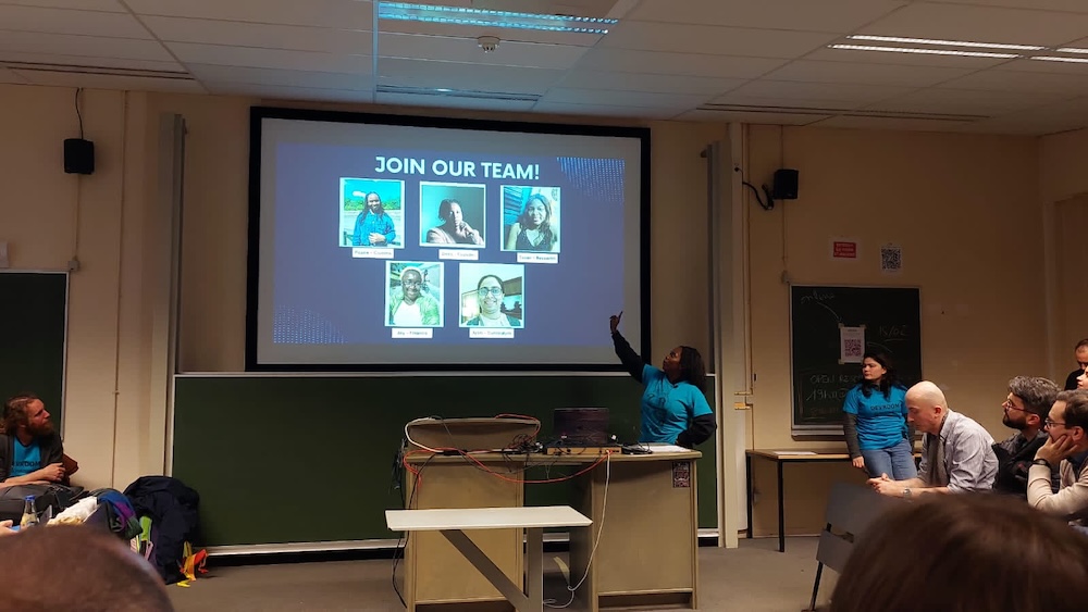 A conference room, with a speaker, Debs, delivering a talk. Debs is an African woman of average height and dark hair. She is wearing a blue &quot;FOSDEM Organisers&quot; t-shirt over her black long-sleeved sweater, and is pointing at the projected slide. The current slide is a blue page with a title in white text, which reads JOIN OUR TEAM with 5 members of an all-female team in display.