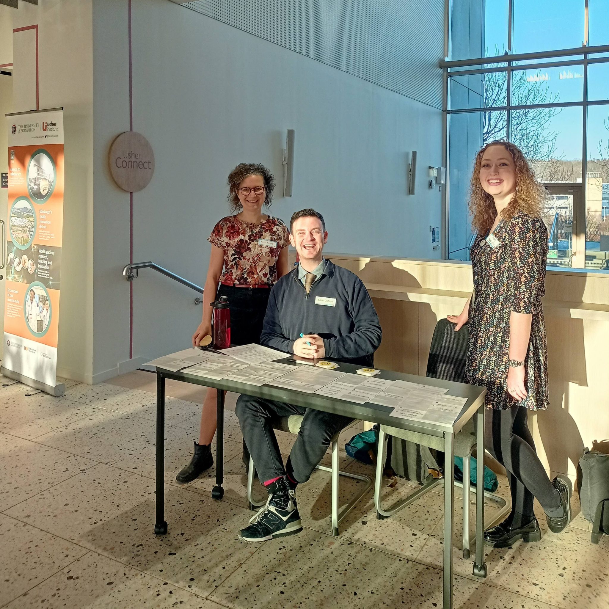 Image of the reception team at the start of the event. Two women are standing behind a desk where a man is sitting. On the desk are name badges. The man and women are all smiling.