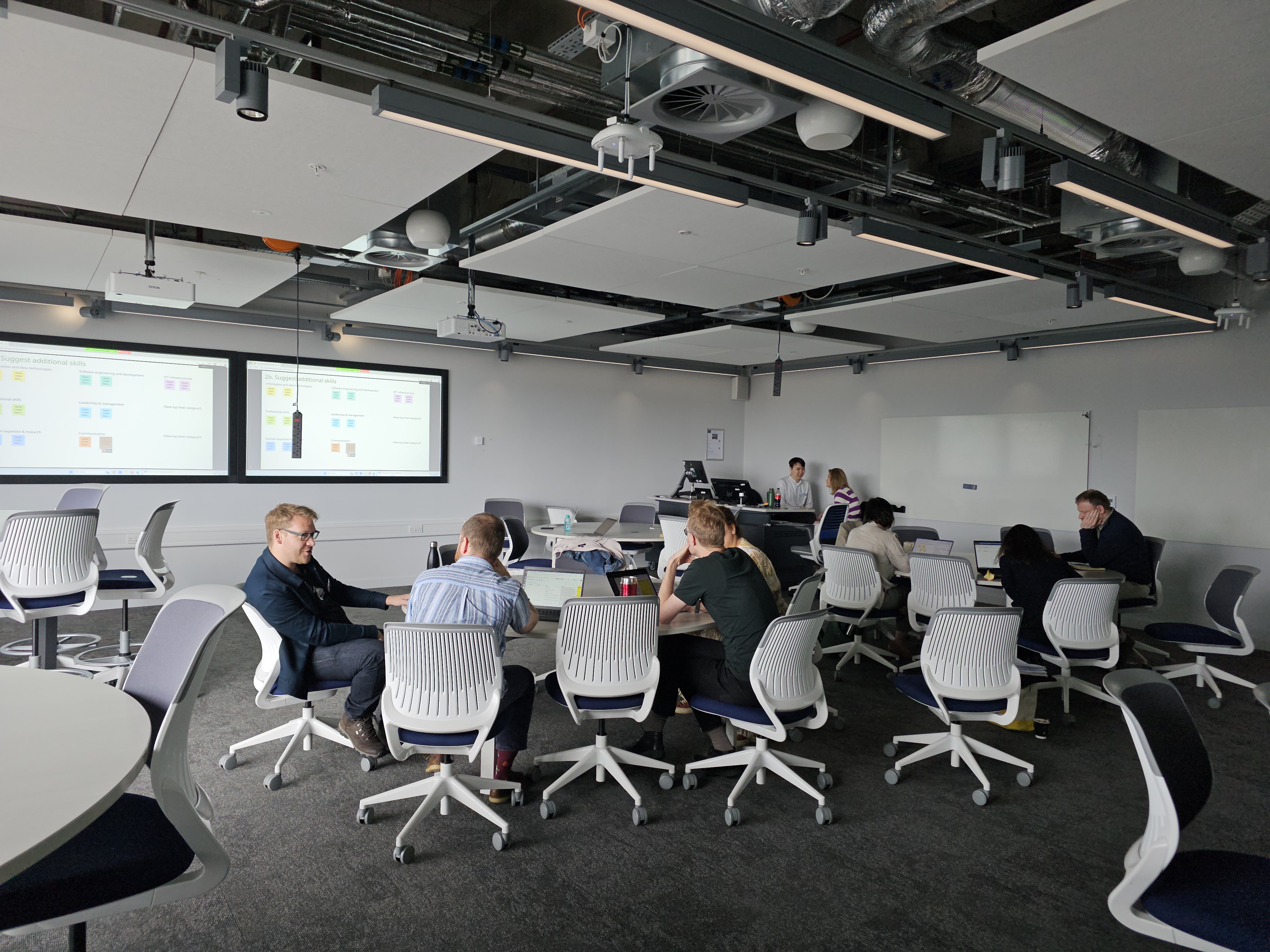 Image of a group of people sitting on chairs around a table and discussing in a meeting room with a screen in the back.